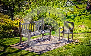 Rustic wooden park bench and chair sit on rock platform on hill in park with large tree shdow and blurred valley behind lush and