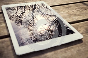 Rustic wooden table with digital tablet and trees reflecting on black screen. View from above