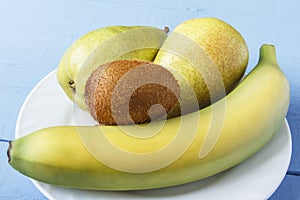 Rustic wooden table with pears, banana and kiwi fruit on white plate. Healthy breakfast or dinner with natural foods. Side view