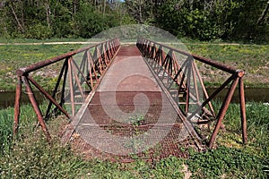 Rusty pedestrian bridge with grate floor as pathway across artificial water channel and triangular shaped railing