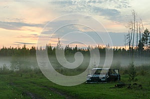 The rusty remains of the Soviet iron car lie in the forest near the road in a fog strip at sunset in the North