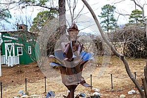 A rusty sculpture of an elf playing a horn in the garden surrounded by bare winter trees, yellow winter grass with gray sky