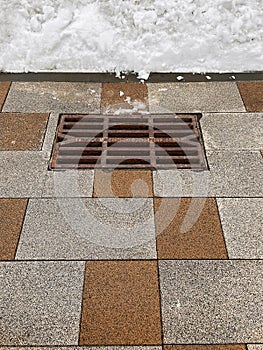 Rusty storm drain grate on tiled road and snowdrift that will melt in spring