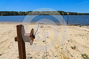 A rusty wench with a long cable in the silky brown sand of the beach on the banks of the rippling lake with lush green treeys