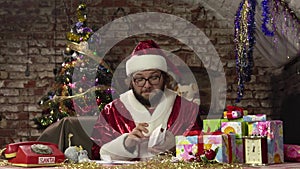 S. Claus is sitting at a table with gifts at home and takes out a children`s letter from an envelope
