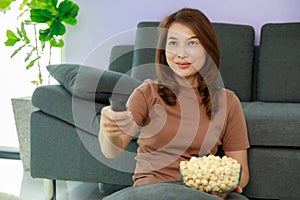 45s cute Asian housewife sitting near sofa and holding remote control and aim it to tolevision with a bowl of popcorn in living