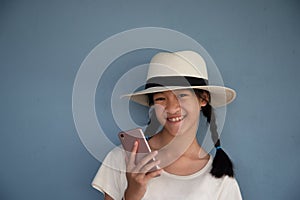 10s smiling long hair Asian girl holding pink mobile phone and wearing hat with blue concrete background