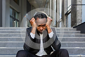 Sad african american businessman sitting on stairs outside office building, man in business suit bankrupt fired from job