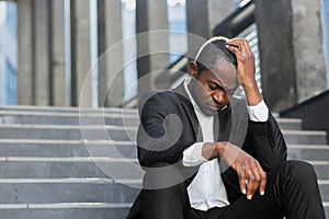 Sad african american businessman sitting on stairs outside office building, man in business suit bankrupt fired from job