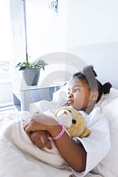 Sad african american girl patient lying on bed with teddy bear in patient room at hospital