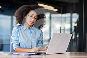 Sad african american remote worker sitting by table and looking desperately in pc screen on blurred background