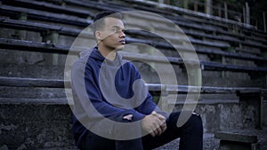 Sad african american teenager sitting on tribune, devastation and poverty around