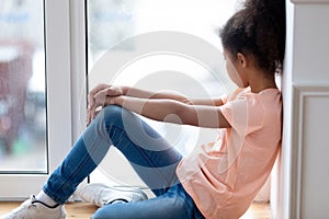 Sad african teenage girl sitting on window sill at home