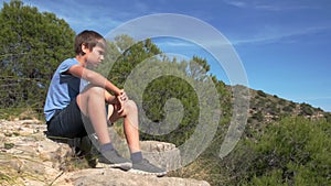 Sad alone boy sitting on the ground in park at sunny day and looking away