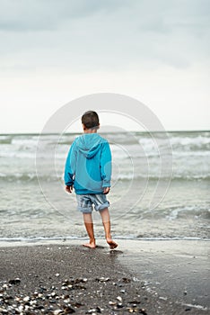 Sad alone kid standing on beach, looking at sea and thinking.