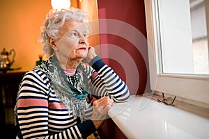 Sad alone senior woman looking through window at home