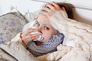 Sad alone young woman in white sweater and blue scarf feeling cold sick and resting home in bed. cleaning her nose, holding her