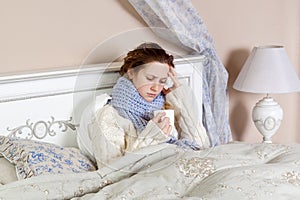 Sad alone young woman in white sweater and blue scarf feeling cold sick and resting home in bed. holding her hot drink and try to