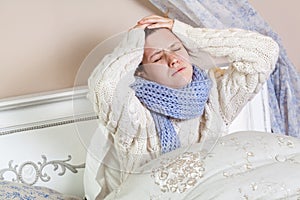 Sad alone young woman in white sweater and blue scarf feeling cold sick and resting home in bed. holding her painful head and