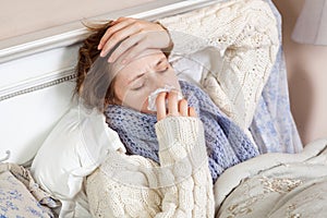 Sad alone young woman in white sweater and blue scarf feeling headache and cold sick and resting home in bed. holding her painful