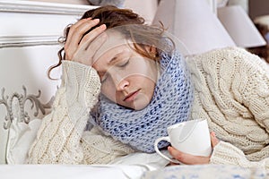 Sad alone young woman in white sweater and blue scarf feeling headache, cold sick and resting home in bed. holding her painful