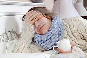 Sad alone young woman in white sweater and blue scarf feeling headache and cold sick and resting home in bed. holding her painful