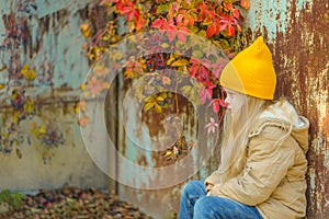 a sad angry little girl sits in the autumn on the street against the background of a peeling wall twined with wild