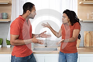 Sad angry young african american guy and lady in same t-shirts quarrel and gesticulate in kitchen interior