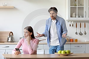 Sad angry young man yelling at woman with smartphone, unhappy lady ignoring him on minimal kitchen