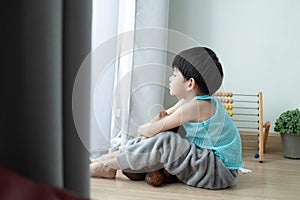 Sad Asian boy sits with his head down and looks out the door at his favorite doll