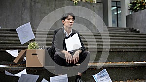 A sad Asian businessman sits on the stairs with a box of his personal stuff after losing his job