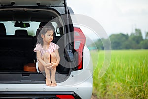 Sad asian child girl sitting alone in a car trunk and looking to the nature outside while going on vacations with their parents.
