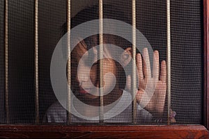 Sad asian child standing behind the wire screen window in dark m