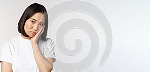 Sad asian girl looking upset and lonely, sulking and frowning, standing against white background in casual tshirt