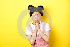 Sad asian girl pleading for help, begging you, sulking and whining, standing on yellow background