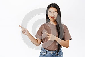 Sad asian girl pointing fingers left, grimacing, making upset face, regret or jealous, white studio background