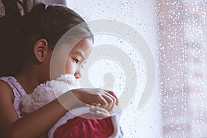 Sad asian little child girl hugging her doll and looking outside