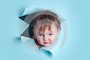 Sad baby in a hole on a paper blue background. Torn child\'s head studi