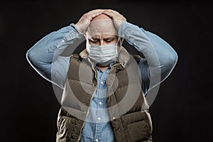 Sad bald adult man in a medical mask stands holding his head with his hands. Precautions quarantined for the period of the