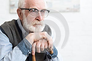 Sad and bearded senior man with walking stick sitting in room