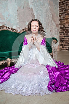 Sad beautiful woman in fantasy white and purple rococo style medieval dress sitting on the floor near sofa and praying