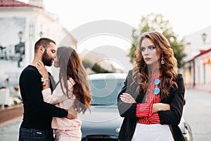 Sad beautiful woman with folded arms in fashionable clothes against kissing couple.