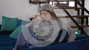 Sad beautiful woman with white wine in glass sitting on sofa thinking. Portrait of depressed young Caucasian lady alone