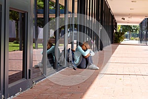 Sad, biracial schoolgirl with school bag sitting on the ground outside school, with copy space