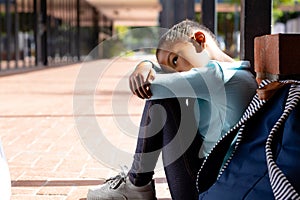 Sad, biracial schoolgirl with schoolbag sitting on the ground outside school in sun with copy space