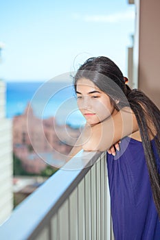 Sad biracial teen girl on outdoor highrise patio, ocean backgro