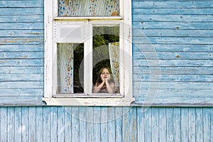 Sad bored little girl looking out the country house window leaning her face on her hands
