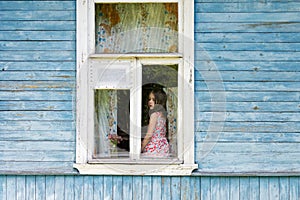 Sad bored little girl sitting on the sill and looking out the country house window