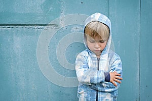 Sad bored resentful child boy in hoodie standing near blue wall. Cute upset distressed kid pouts his lips. Emotional portrait