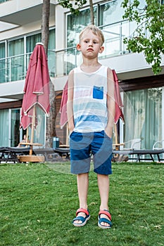 Sad boy in beach clothes stands on a well-maintained lawn on background of modern seaside hotel